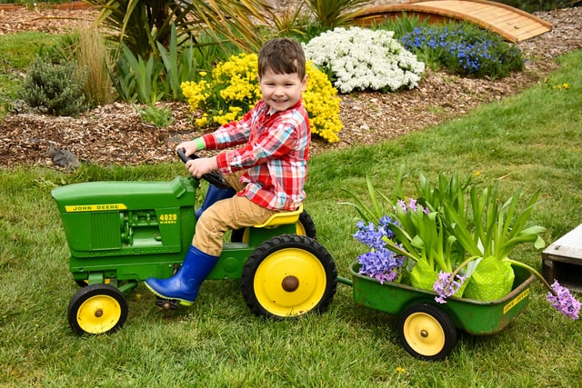 young boy wearing tan jeans and a red plaid shirt riding a green tractor with a trailer carrying plants in a green yard